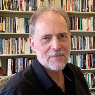 Bruce Bimber smiles at the camera in a black collared shirt with a bookcase behind him