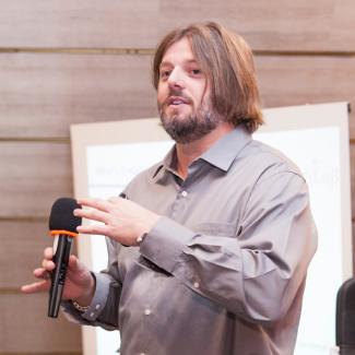 Homero Gil de Zuñiga speaks holding a microphone and wearing a grey shirt