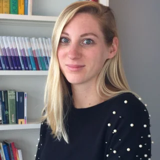 Nadine Bol smiles wearing a blue sweater with beaded sleeves in front of a bookcase