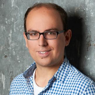 Stephan Winter smiles and poses against a cement wall wearing a blue and white collared checkered shirt