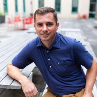 David DeAndrea smiles for the camera while sitting at a picnic table outside in a blue shirt