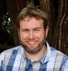 Joshua Meyer-Gutbrod smiles wearing a blue and white plaid shirt