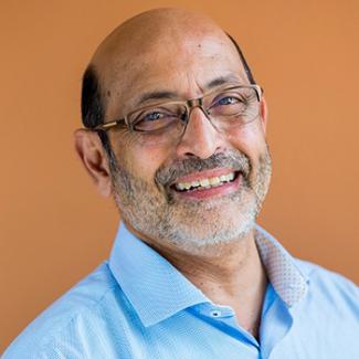 Umesh Mishra smiles for a portrait, wearing glasses and a blue shirt in front of an orange background