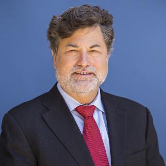 Charles Hale smiles for a portrait photograph wearing a red tie and black blazer in front of a blue background